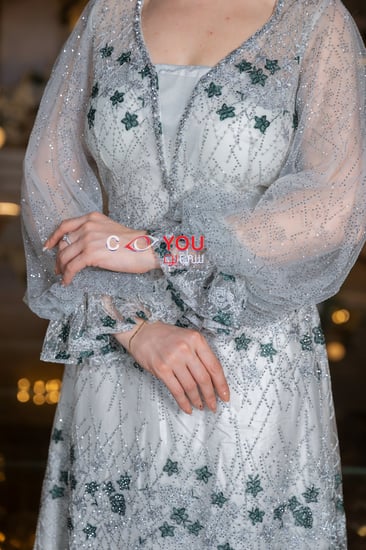 فستان سهرة أبيض مطرز بزهور خضراء أنيقة مع أكمام شفافة منفوشة بتصميم فاخر، لإطلالة ملكية في حفلات الزواج والمناسبات المميزة.