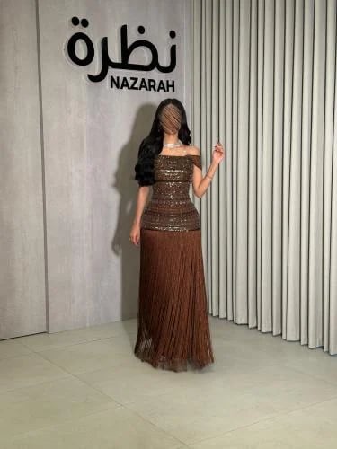 فستان سهرة بقصة أوف شولدر، مزين بتطريزات من الخرز والترتر على الصدر والخصر، مع هدب طويل من الأسفل