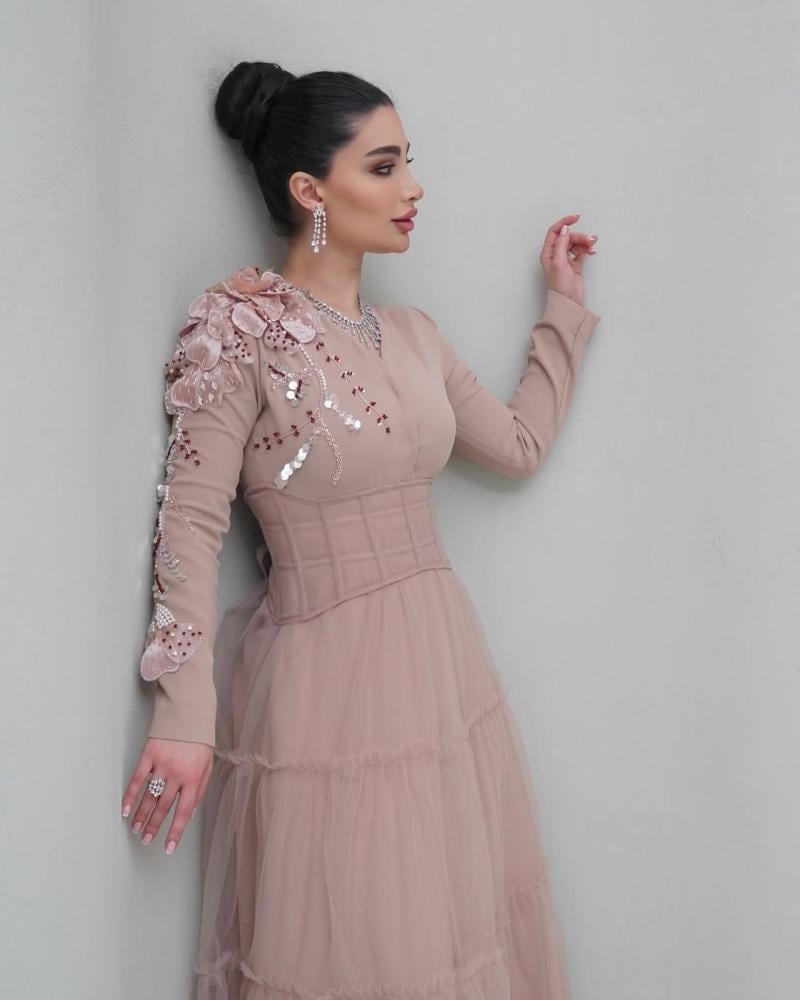 Elegant and soft dress in a nude color with a pinkish tint, decorated with embroidery on the shoulder, long sleeves, with a wide belt at the waist to shape the body, made of Barbie fabric and tulle from Jawhara store