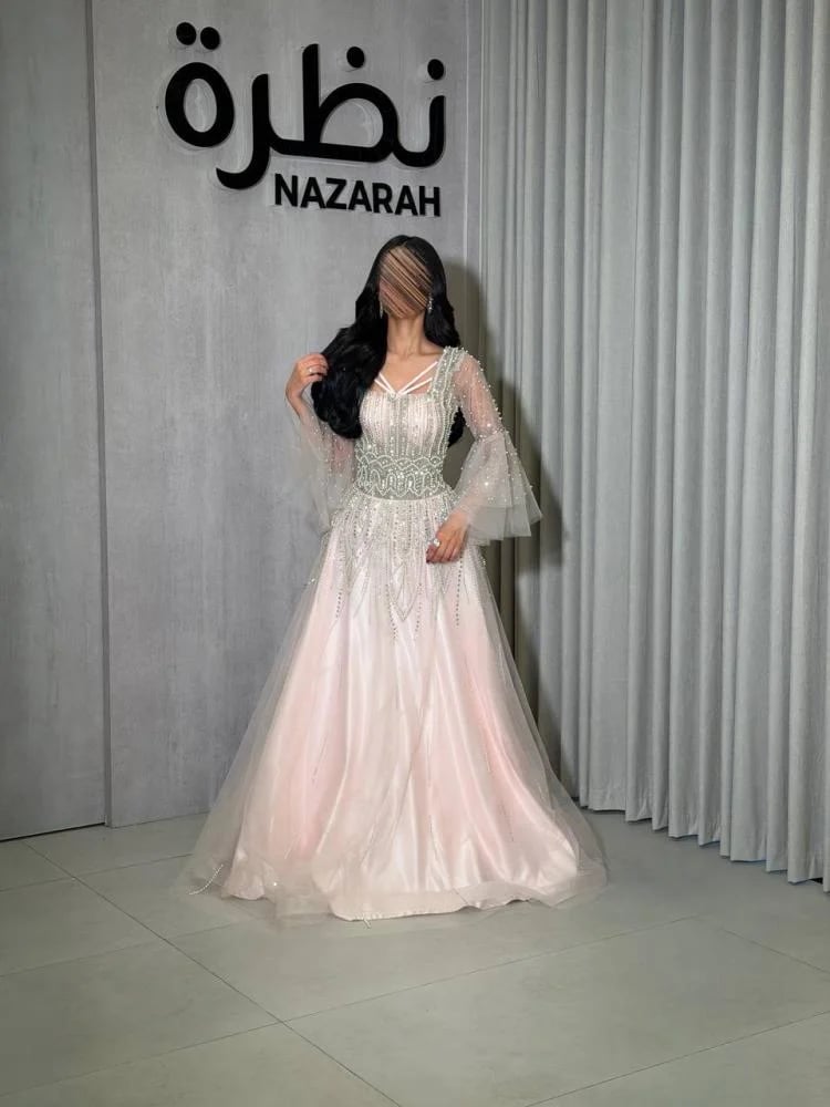 فستان سهرة بقصة كورسيه مطرز بالخرز واللؤلؤ، بأكمام شفافة واسعة من التول، وتفاصيل تطريز ممتدة على كامل الفستان.