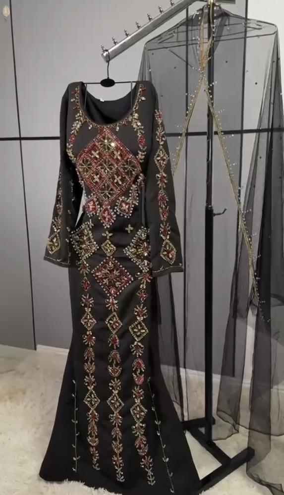 مخور أسود من قماش كريب ناعم، مزين بتطريز يدوي وأحجار لامعة بالأحمر والذهبي، مع رداء تول شفاف طويل يمنحه إطلالة ملكية أنيقة ومميزة.