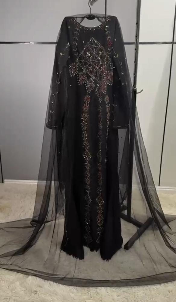 مخور أسود من قماش كريب ناعم، مزين بتطريز يدوي وأحجار لامعة بالأحمر والذهبي، مع رداء تول شفاف طويل يمنحه إطلالة ملكية أنيقة ومميزة.